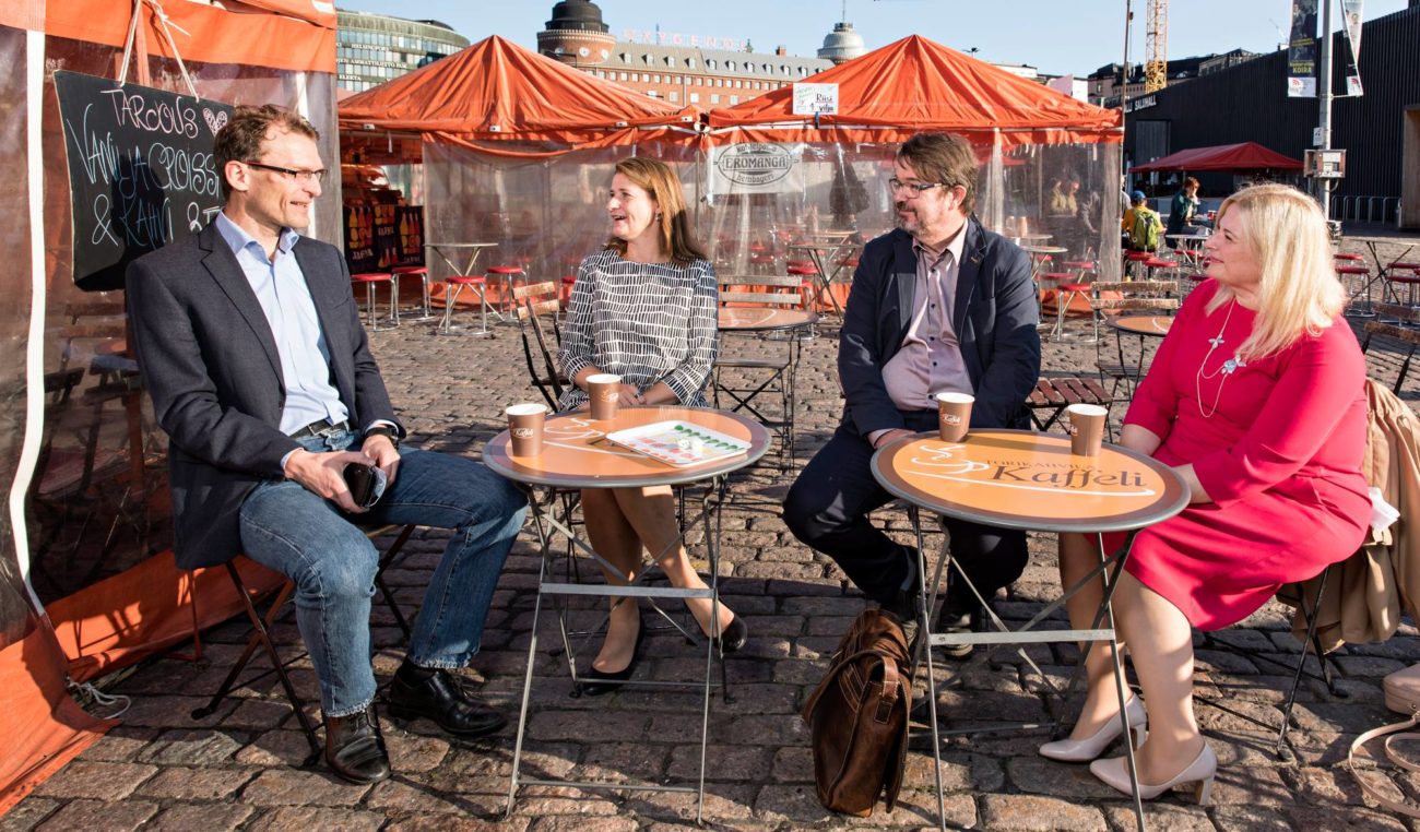 Neljä henkilöä keskustelevat Hakaniemen torilla kahvikojun pöytien ääressä.