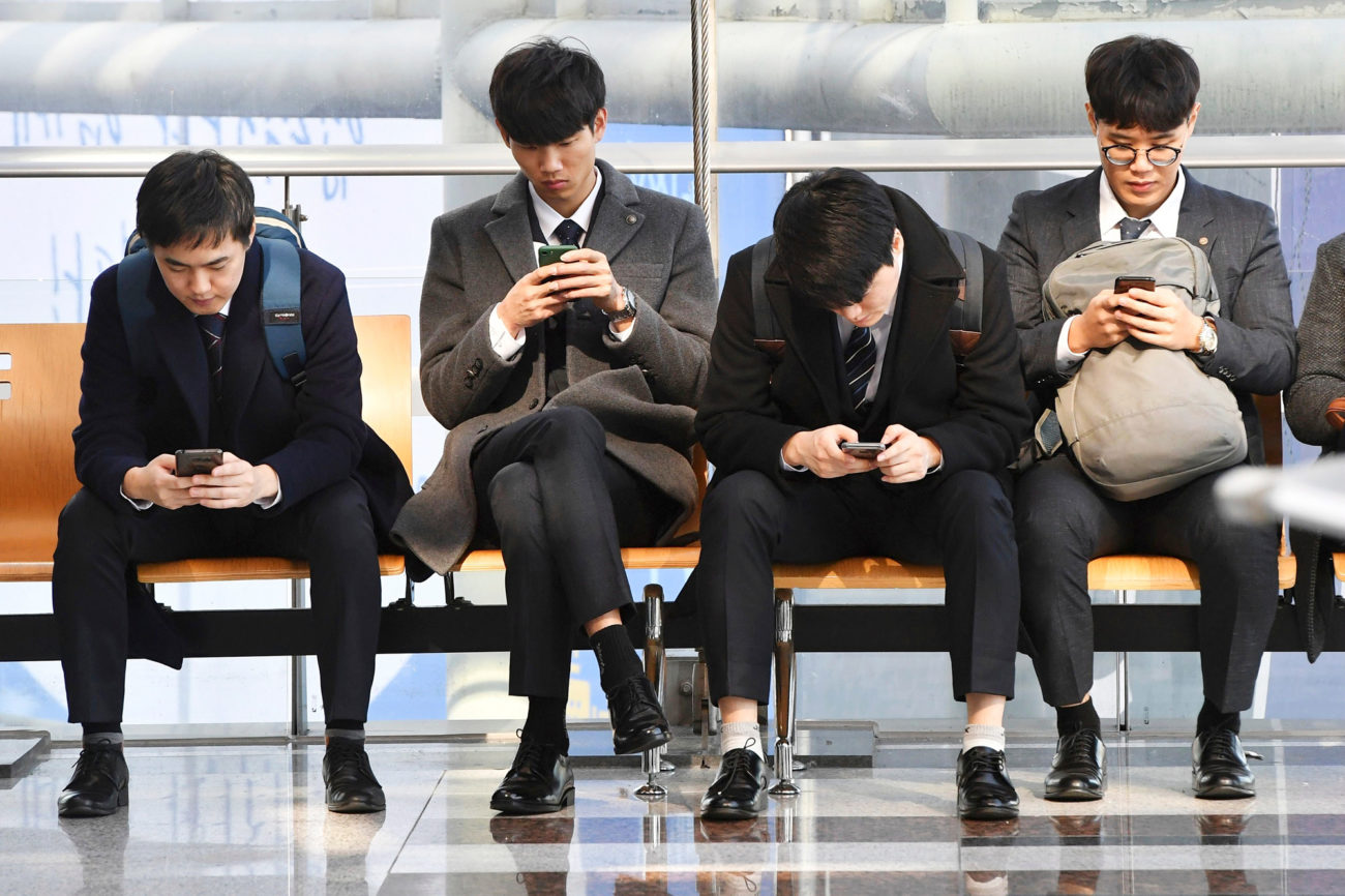 Neljä nuorta korealaismiestä päällysvaatteissaan istumassa odotusaulassa,. Jokainen heistä on pää alas päin syventyneenä matkapuhelimen näpelöimiseen.