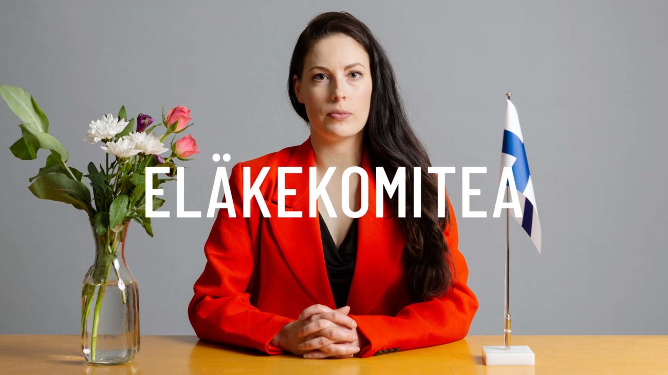 Nainen istuu puheenjohtajan paikalla pöydän ääressä, kädet ristissä katsoo kohti lukijaa. Henkilön virellä pöydällä pöytälippuna Suomen lippu ja maljakossa kukkia.