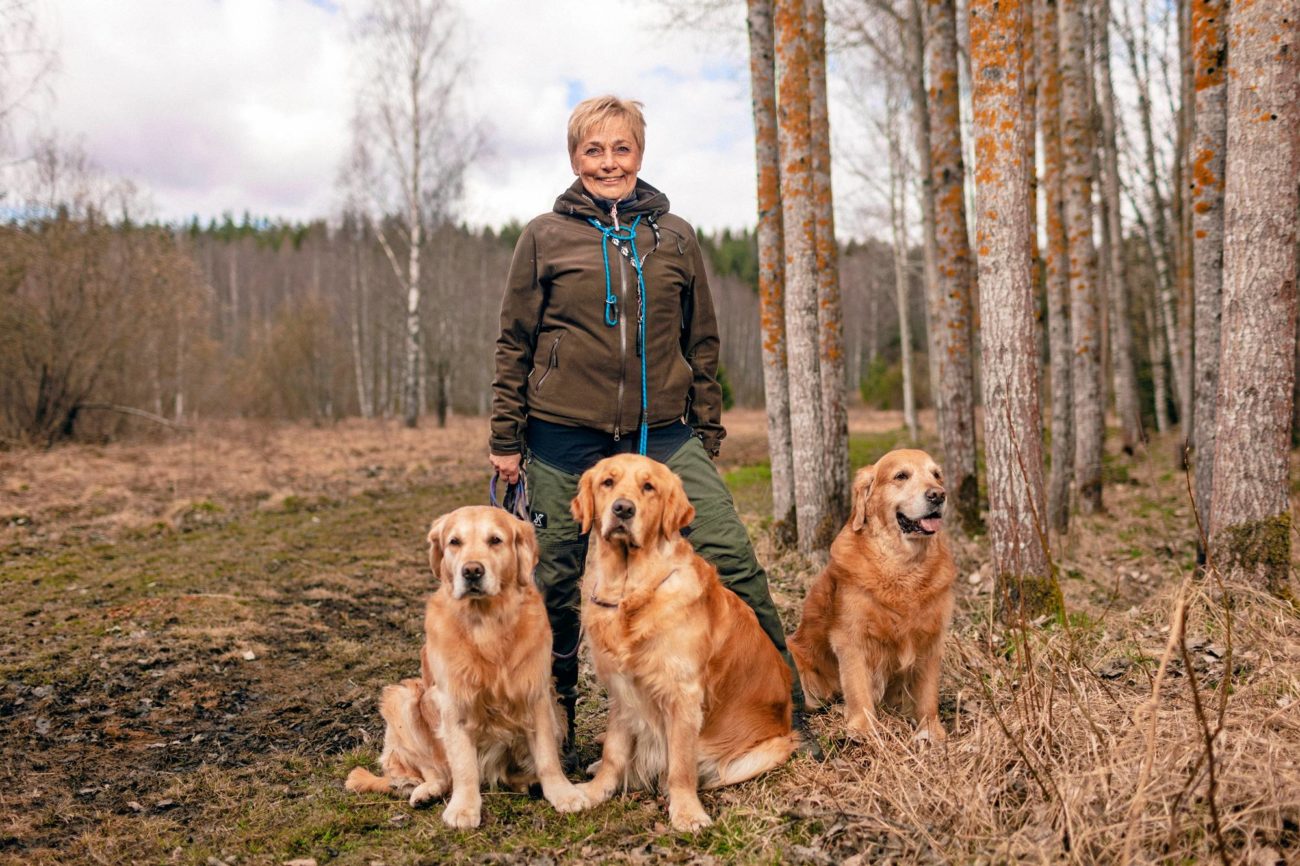 Nainen ja kolme koiraa kuvattuna luonnossa, peltoa ja metsää taustalla.